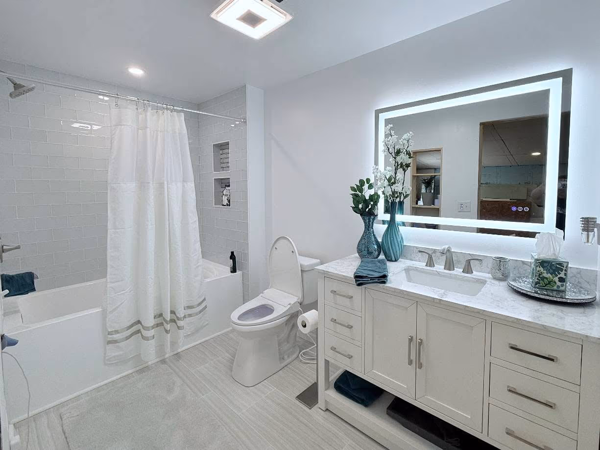 Finished bright bathroom with a white double vanity, illuminated mirror, bathtub, and large-format floor tile